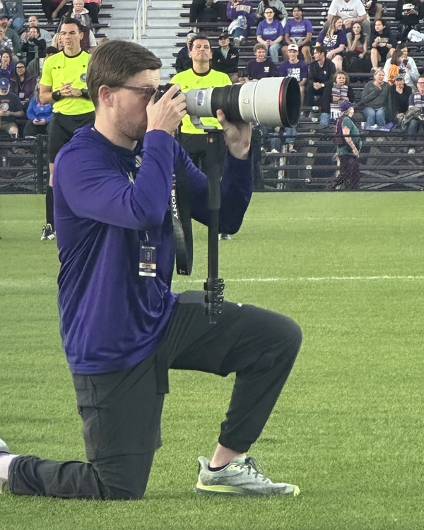 Adam Bender kneeling on the sideline with a camera at a Washington soccer match